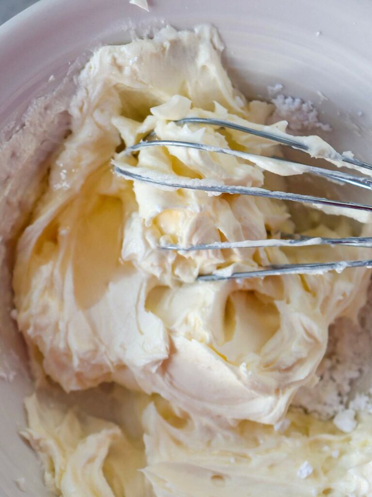 A whisk mixing creamy butter and powdered sugar in a white bowl.