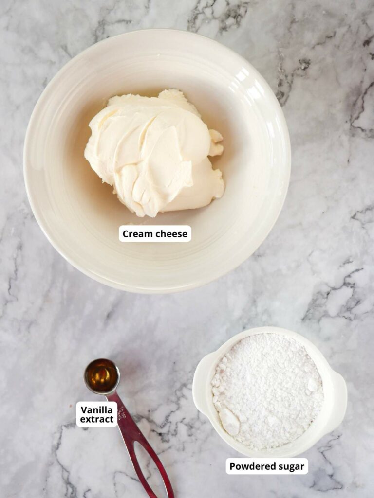 Bowl of cream cheese, a measuring spoon with vanilla extract, and a bowl of powdered sugar on a marble surface.