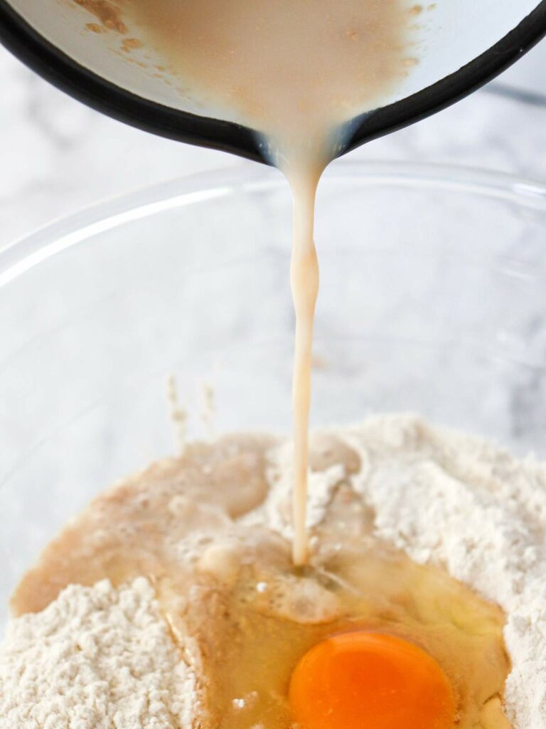 Liquid being poured into a mixing bowl containing flour and a cracked egg.