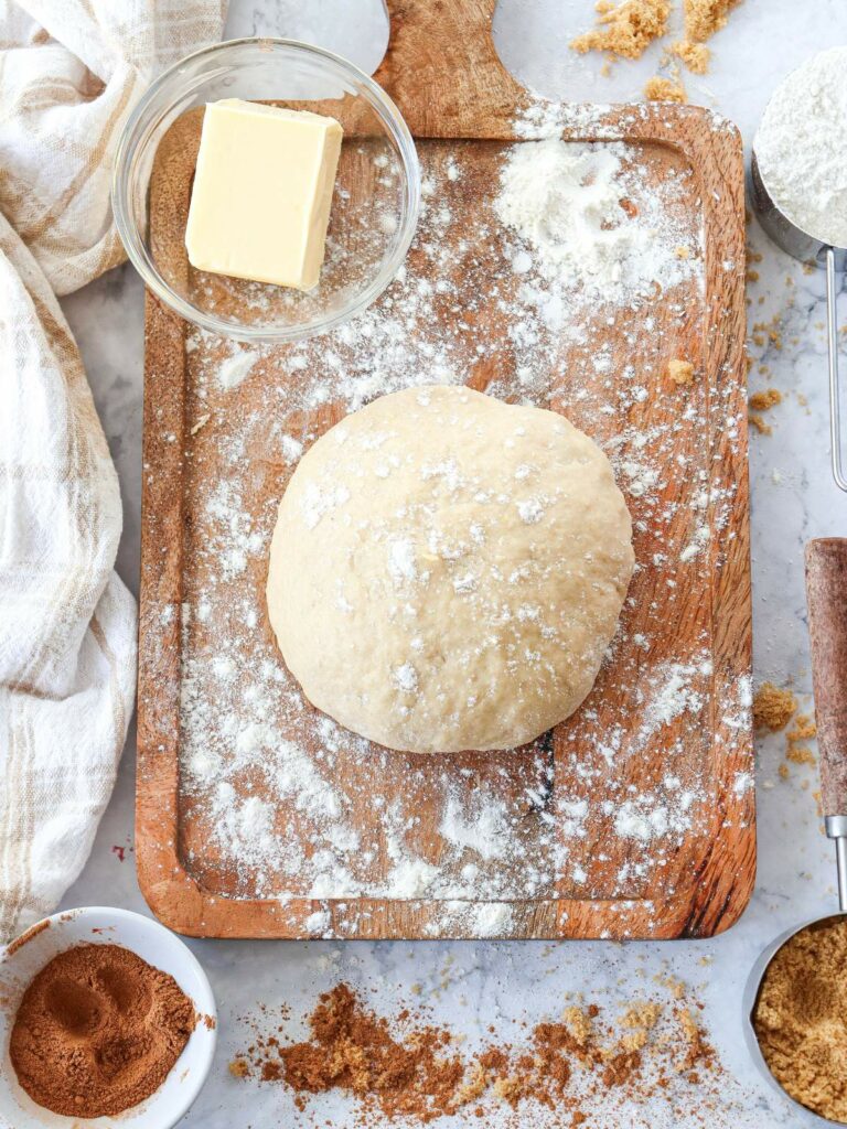 Ball of dough on a floured wooden board, surrounded by a bowl of butter, flour, brown sugar, and cinnamon.