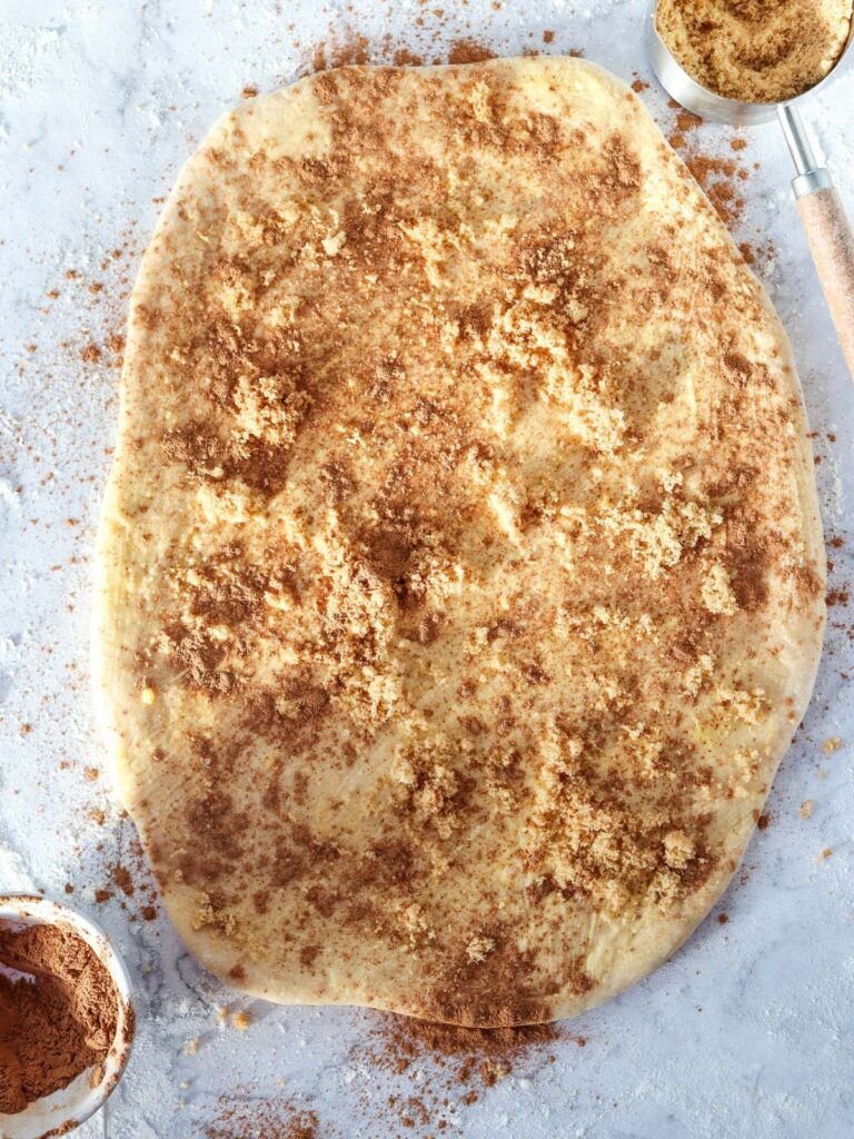 Rolled-out dough covered with cinnamon and sugar on a floured surface, with bowls of cinnamon and sugar nearby.