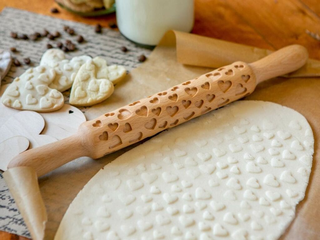 Rolling pin with heart patterns embossing dough; heart-shaped cookies and parchment paper beside it.