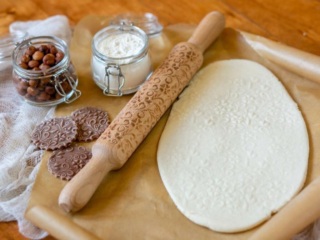 Embossed rolling pin with patterned dough on parchment paper, alongside a jar of flour, a jar of hazelnuts, and patterned cookies.