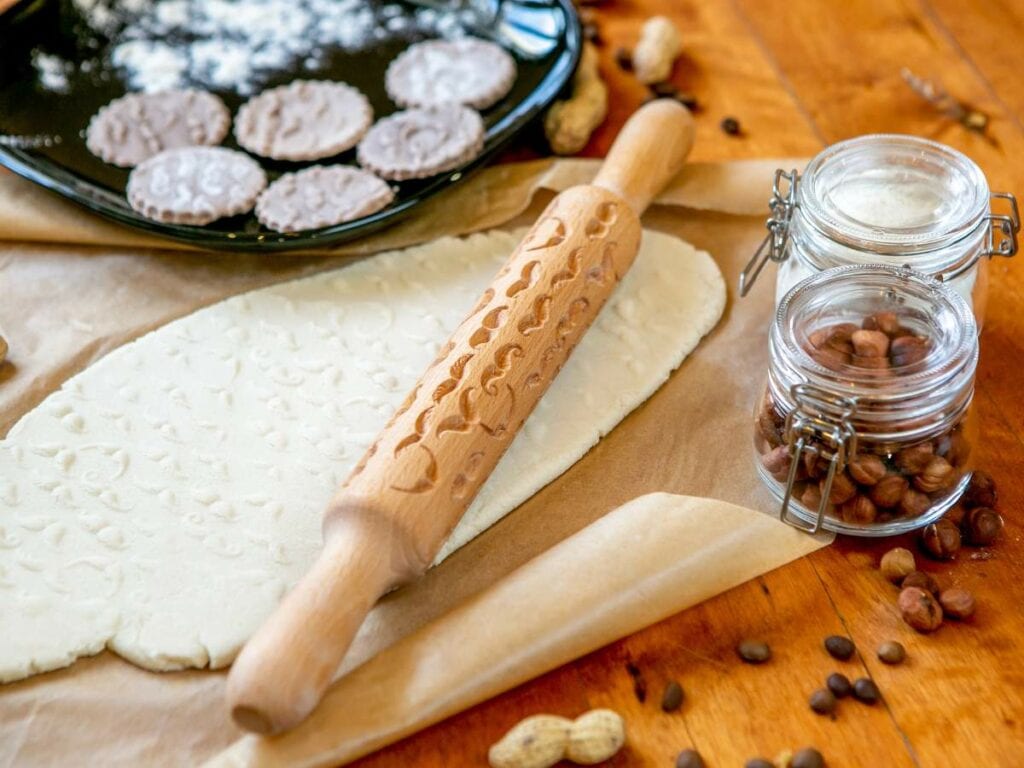 Embossed rolling pin on rolled pastry dough, surrounded by a jar of nuts, scattered peanuts, coffee beans, and cookies on a wooden table.