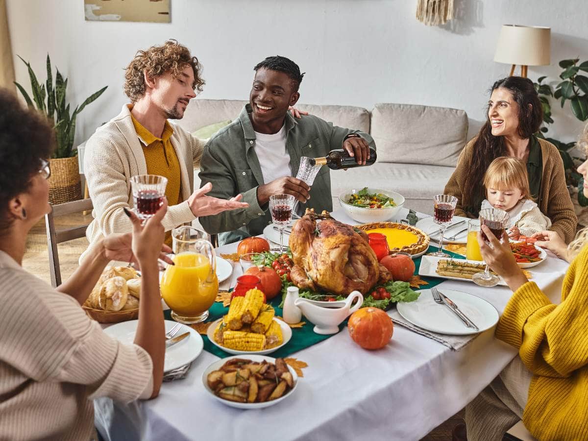 Group of people enjoying a meal around a table with a roasted turkey, corn, salad, and drinks in a cozy dining room.