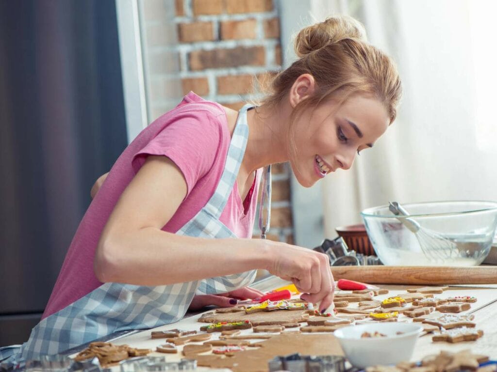 A woman in a pink shirt and apron decorates cookies with icing in a kitchen setting.