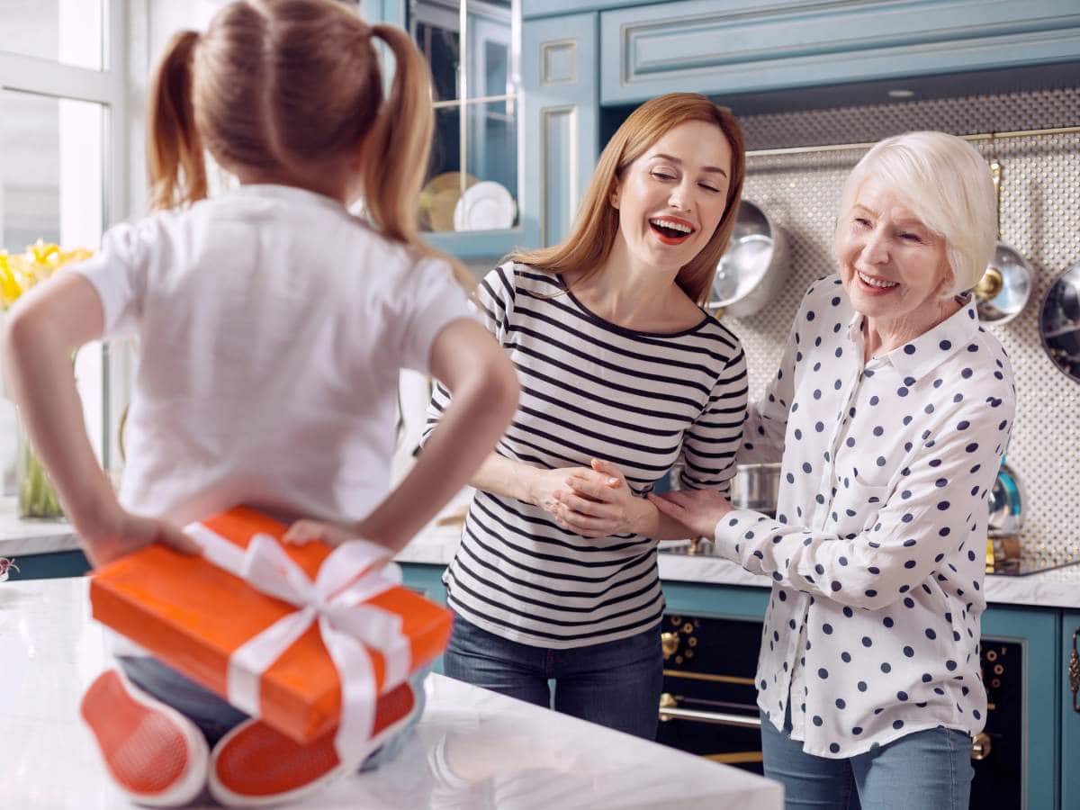 A child with a gift box stands on a kitchen counter, facing two smiling women.