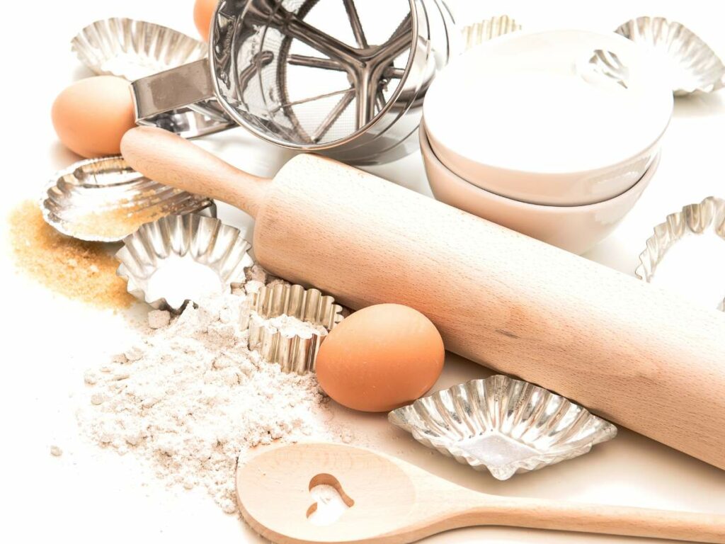 Baking tools and ingredients including a rolling pin, eggs, flour, sugar, a sifter, and baking molds are arranged on a white surface.