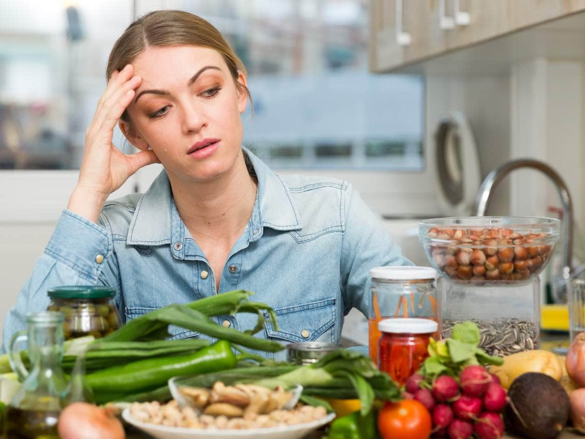 A woman in a kitchen looks stressed, surrounded by various fresh vegetables, nuts, and jars.