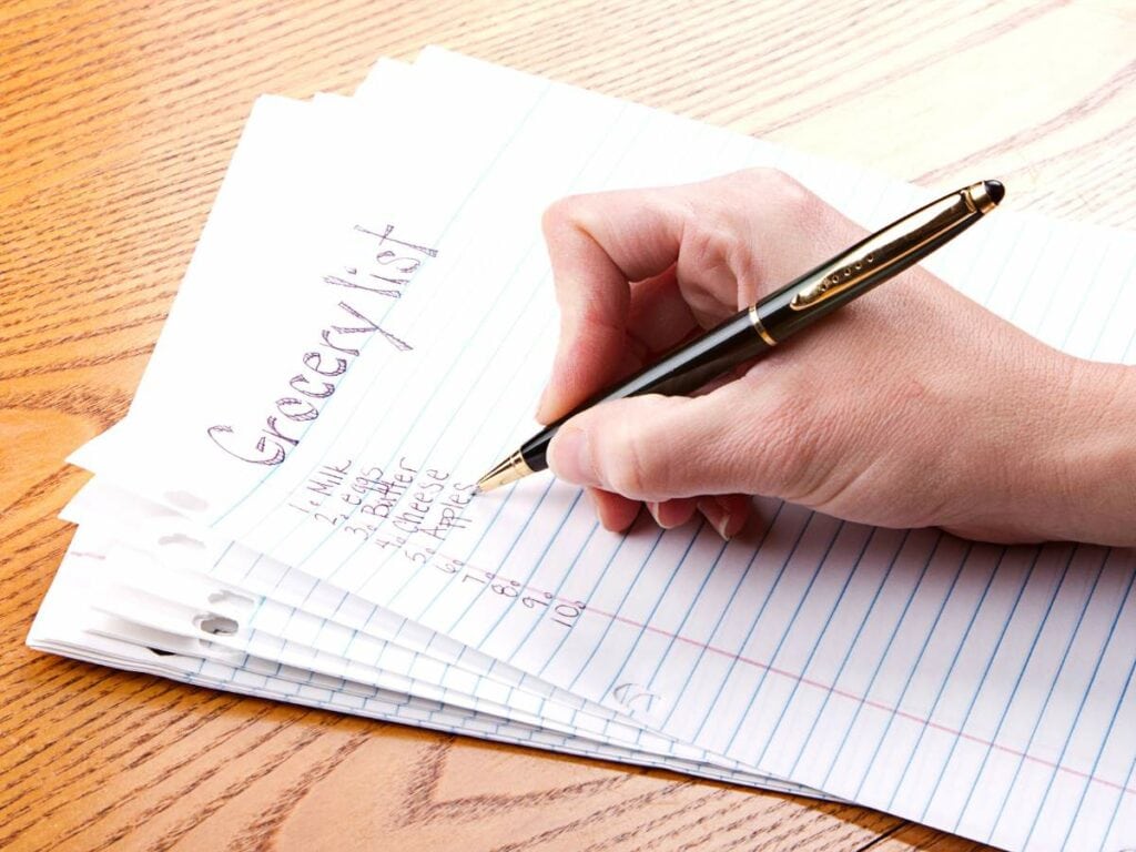 Hand holding a pen writing a grocery list on lined paper, with the words "Grocery List" and items like milk, eggs, and bread visible.