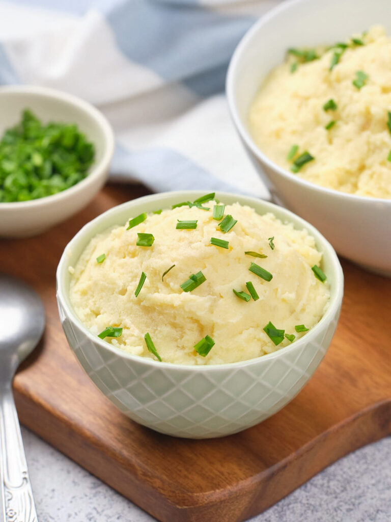 Bowl of mashed potatoes garnished with chopped green onions, placed on a wooden board.