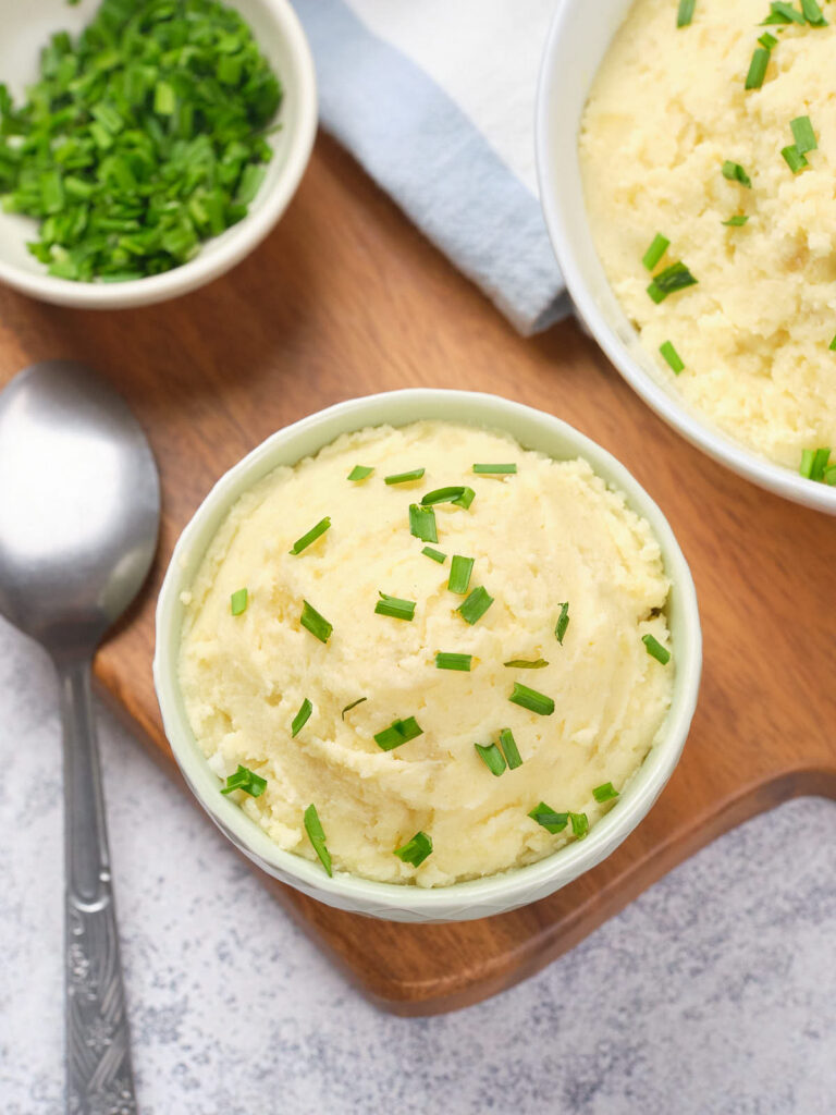 Bowl of mashed potatoes topped with chopped green onions on a wooden board.