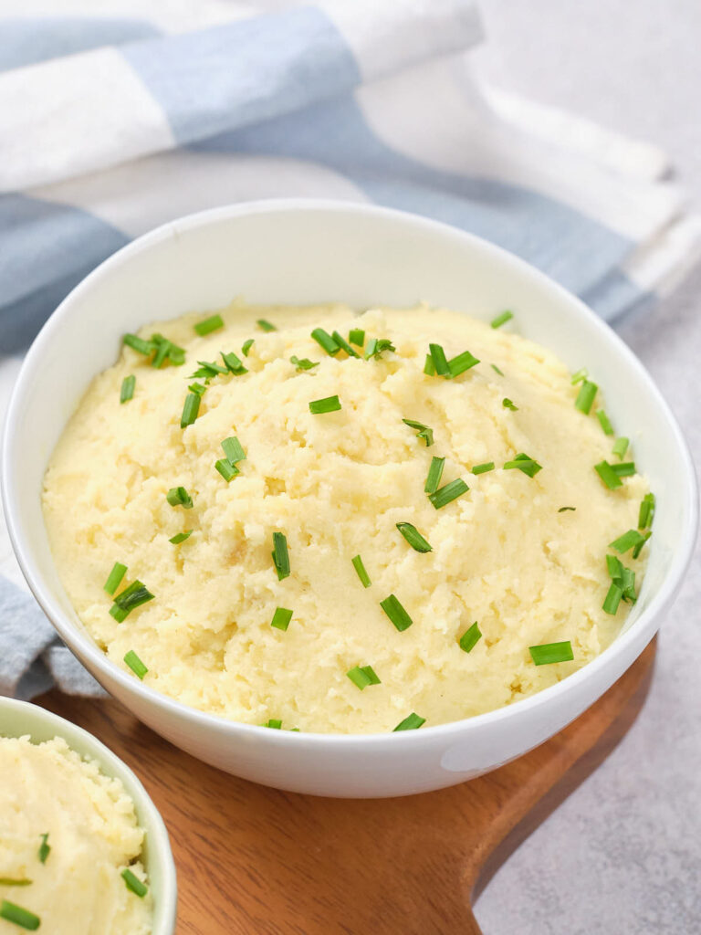 A bowl of creamy mashed potatoes topped with chopped green onions on a wooden surface.