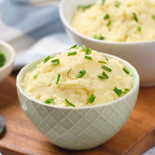 Two bowls of mashed potatoes garnished with chopped chives on a wooden board.