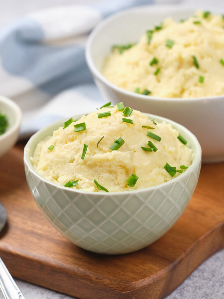 Two bowls of mashed potatoes garnished with chopped chives on a wooden board.