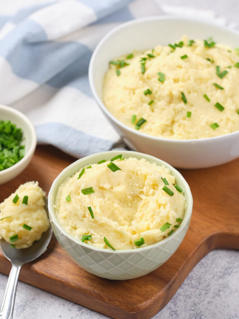 Two bowls of mashed potatoes garnished with chopped chives on a wooden board, next to a small bowl of extra chives.