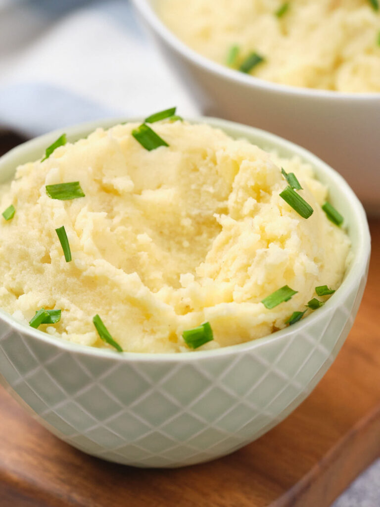 A bowl of creamy mashed potatoes garnished with chopped chives, served in a patterned bowl on a wooden surface.