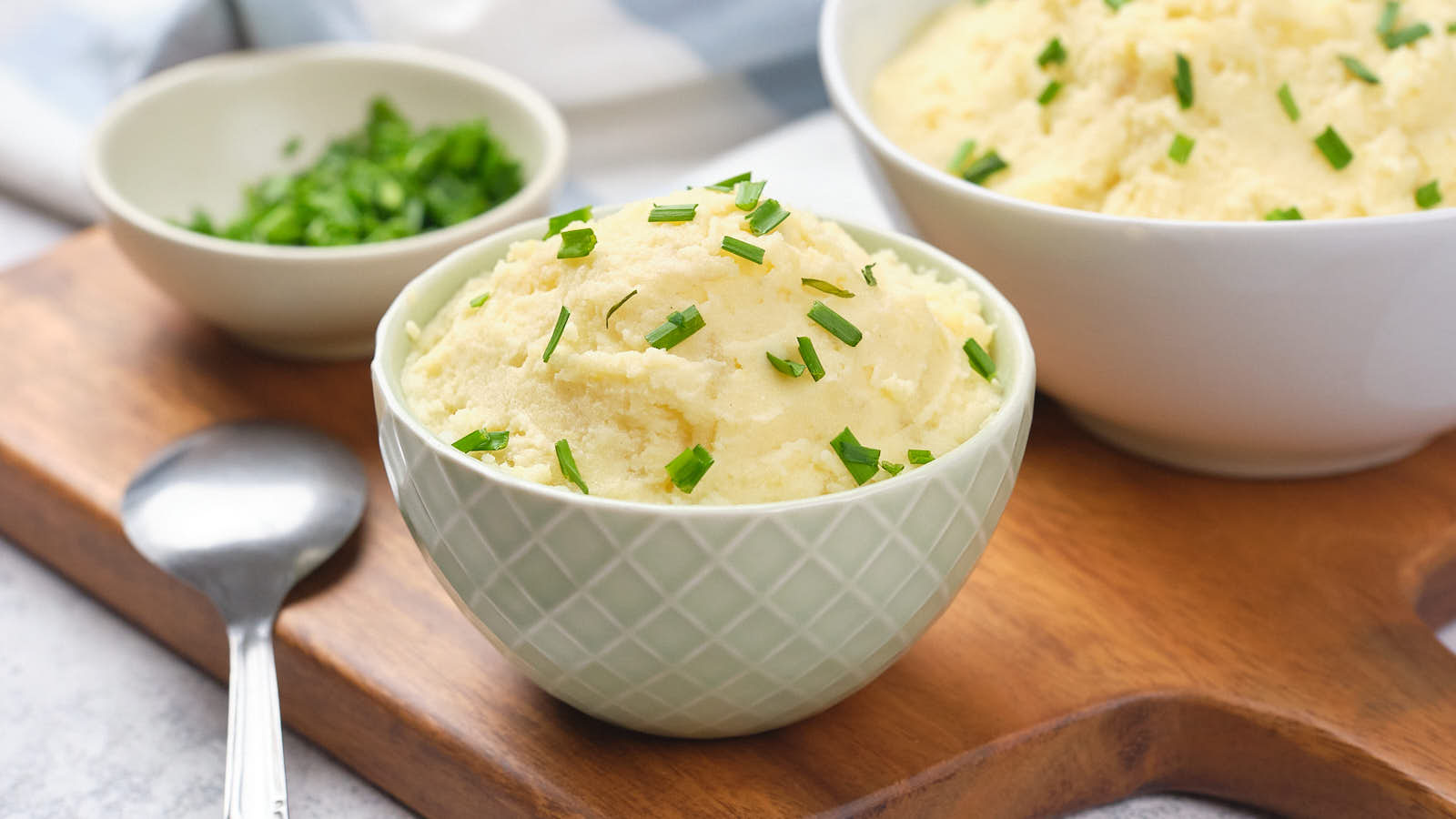 A bowl of mashed potatoes with chopped chives on a wooden board, accompanied by a spoon and a small dish of chives.