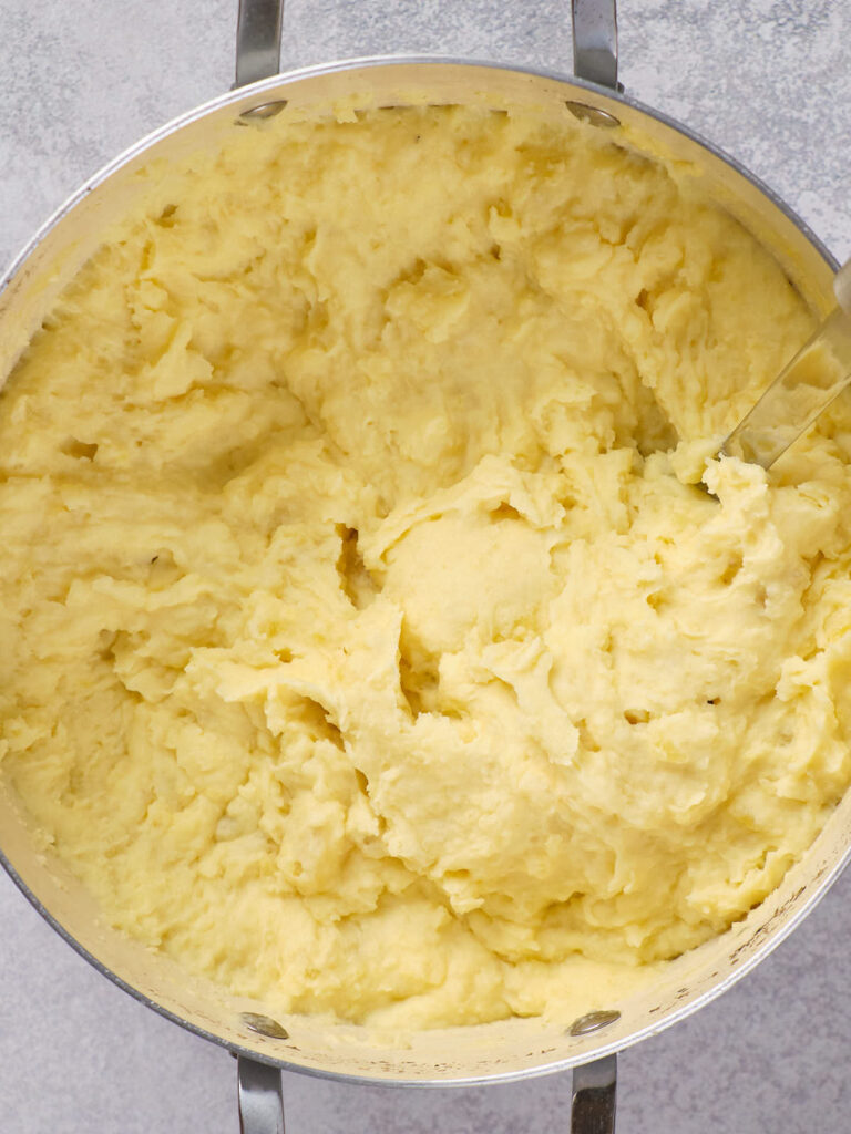 Mashed potatoes in a large pot with a metal spoon on a gray surface.