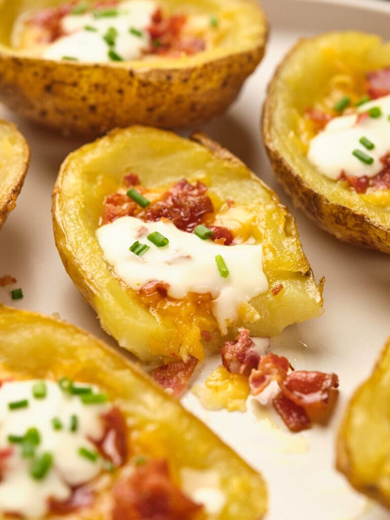 Close-up of potato skins topped with melted cheese, bacon bits, sour cream, and chopped chives on a white plate.