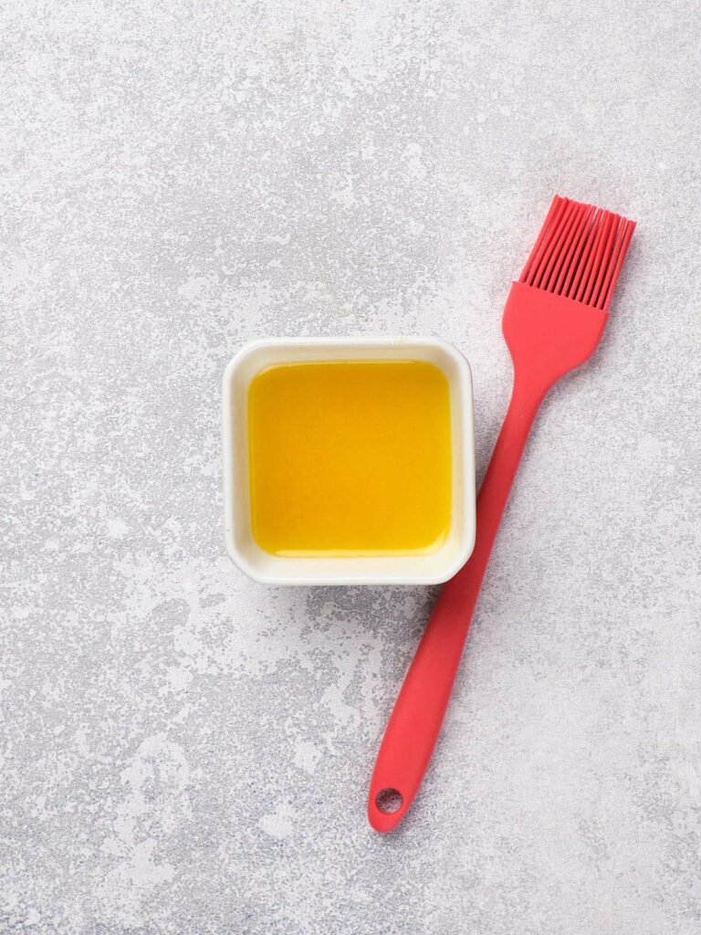 A red silicone brush beside a square white container filled with melted butter on a textured gray surface.