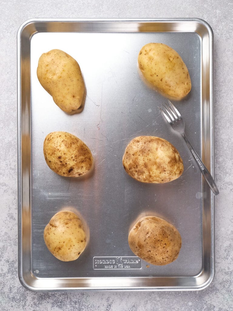 Six unpeeled potatoes are placed on a metal baking sheet with a fork resting beside them.