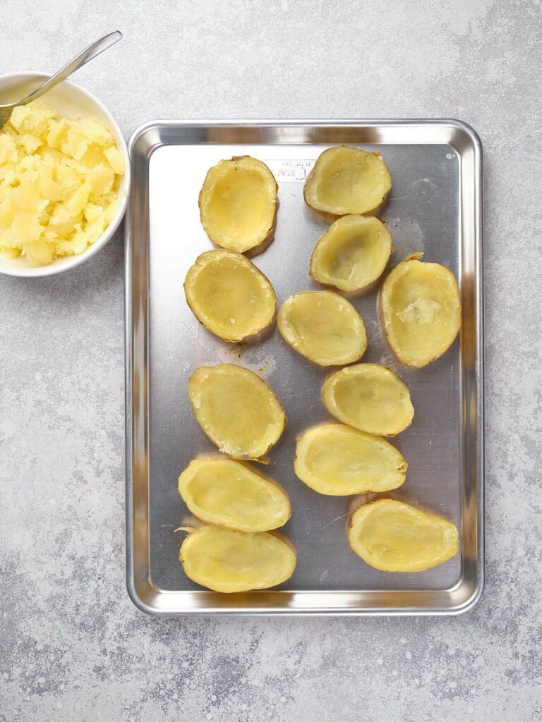 Potato skins arranged on a baking tray with a bowl of scooped potato filling beside them.