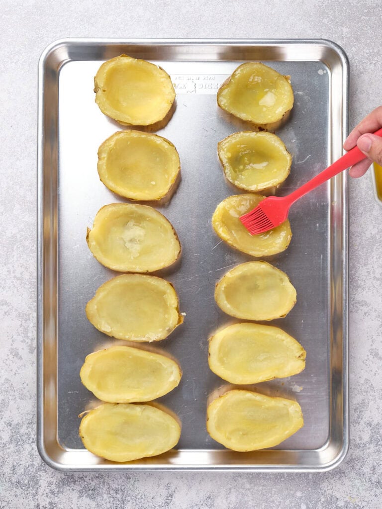 Hands brushing butter on hollowed potato halves arranged on a baking sheet.