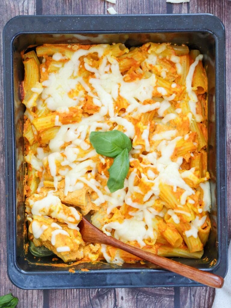 Pumpkin pasta covered in melted cheese, served in a rectangular pan with a wooden spoon, garnished with a basil leaf.