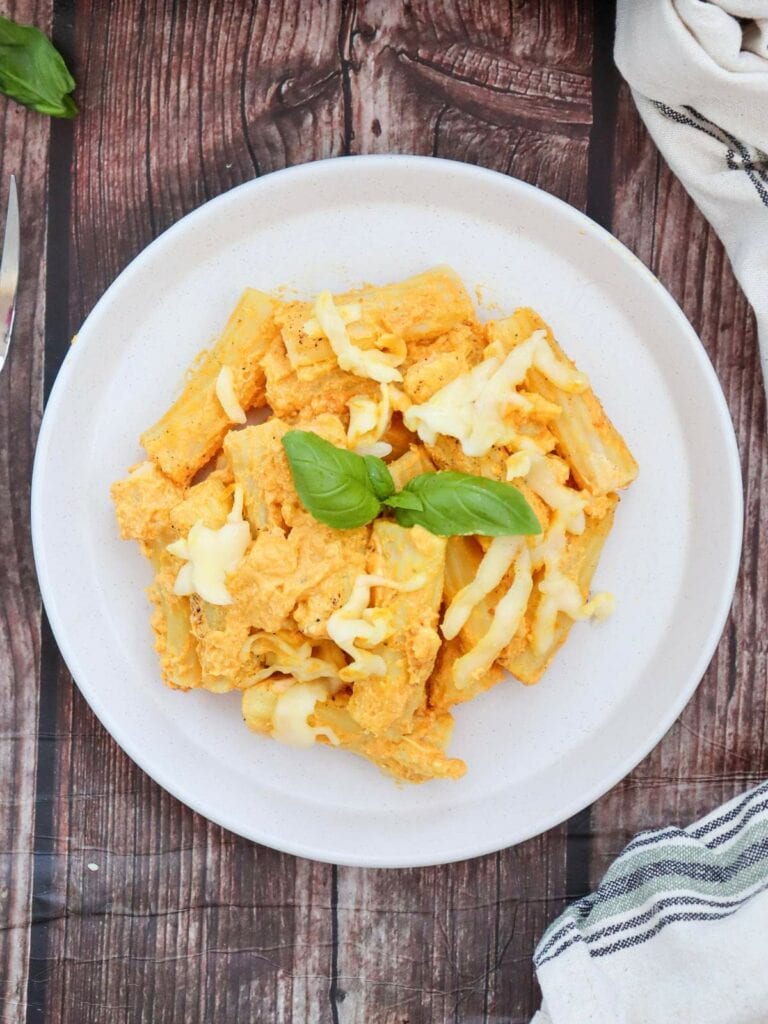 A plate of pumpkin pasta in a creamy orange sauce, topped with melted cheese and garnished with fresh basil leaves, on a wooden table.