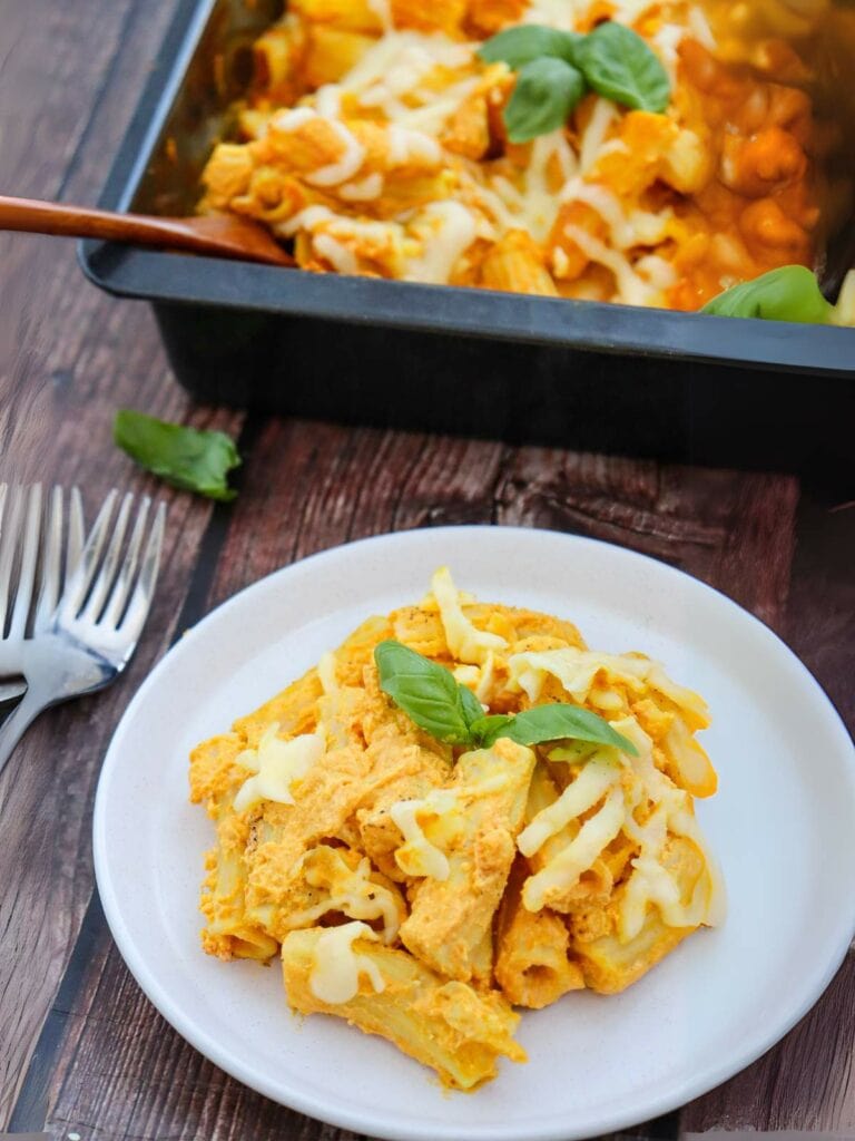 A plate of pumpkin pasta with cheese and basil leaves in front of a baking dish on a wooden table. Two forks are nearby.