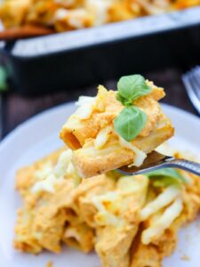 A fork holds a piece of pumpkin pasta garnished with cheese and fresh basil, with a plate of similar pasta in the background.