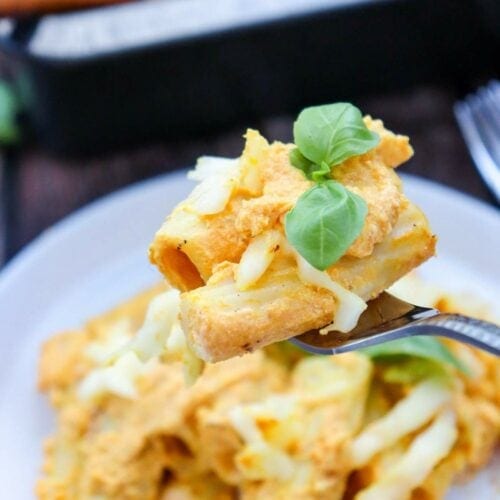 A fork holds a piece of pumpkin pasta garnished with cheese and fresh basil, with a plate of similar pasta in the background.