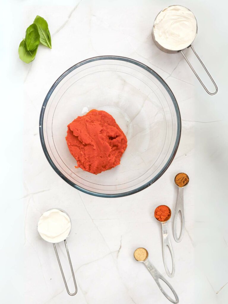 A bowl with pumpkin puree, two measuring cups with cream, and three measuring spoons with spices on a white marble surface.