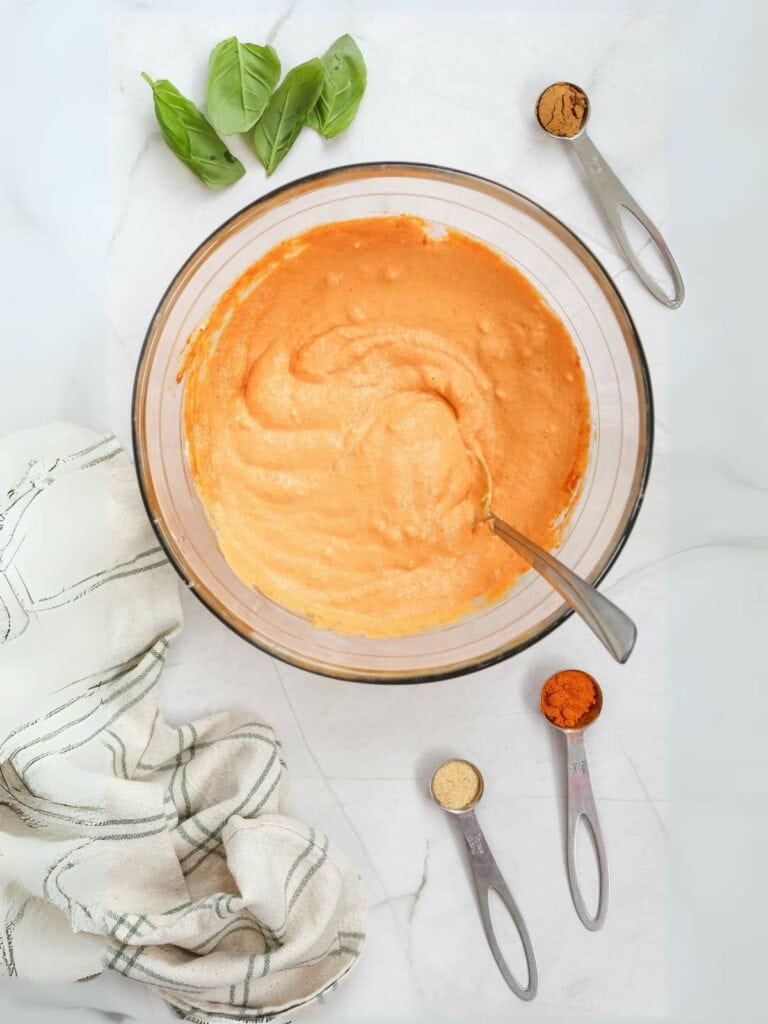 A bowl of orange batter with a spoon, surrounded by basil leaves, measuring spoons with spices.