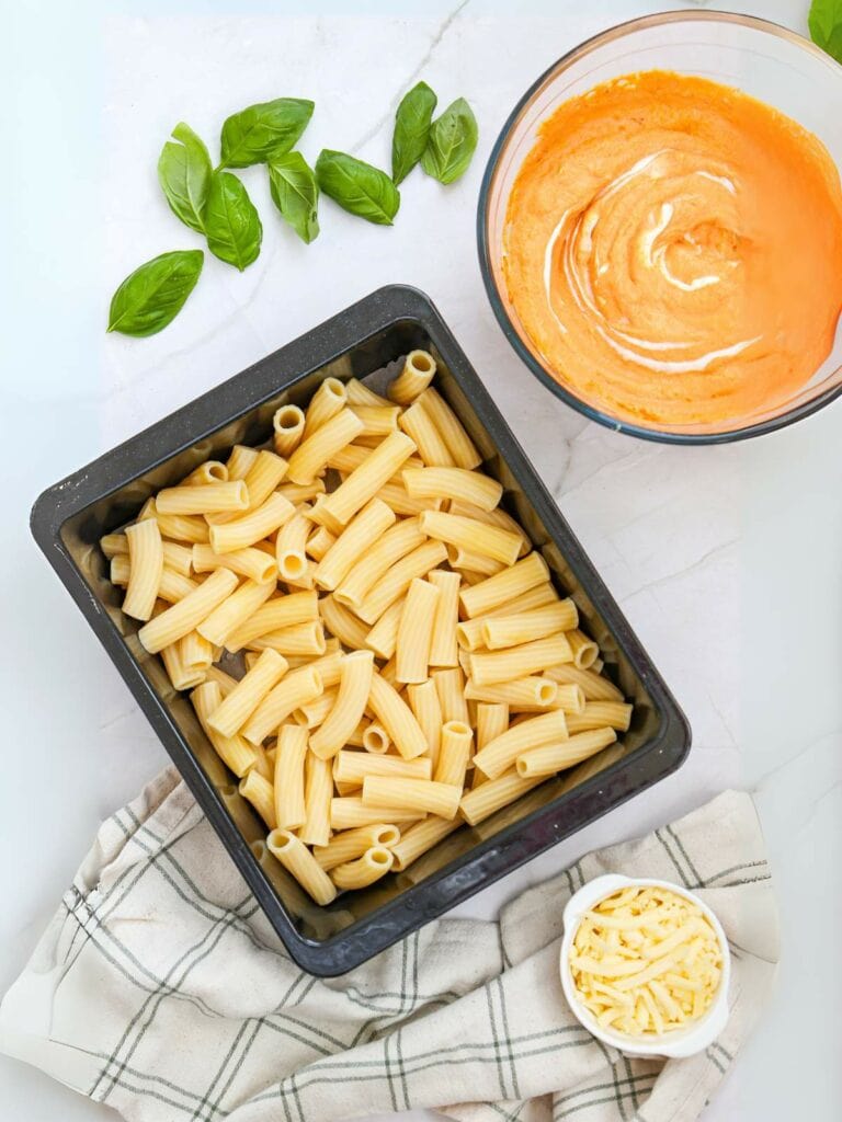 Uncooked rigatoni in a black tray, creamy tomato sauce in a glass bowl, fresh basil leaves, shredded cheese in a small bowl, and a checked cloth.