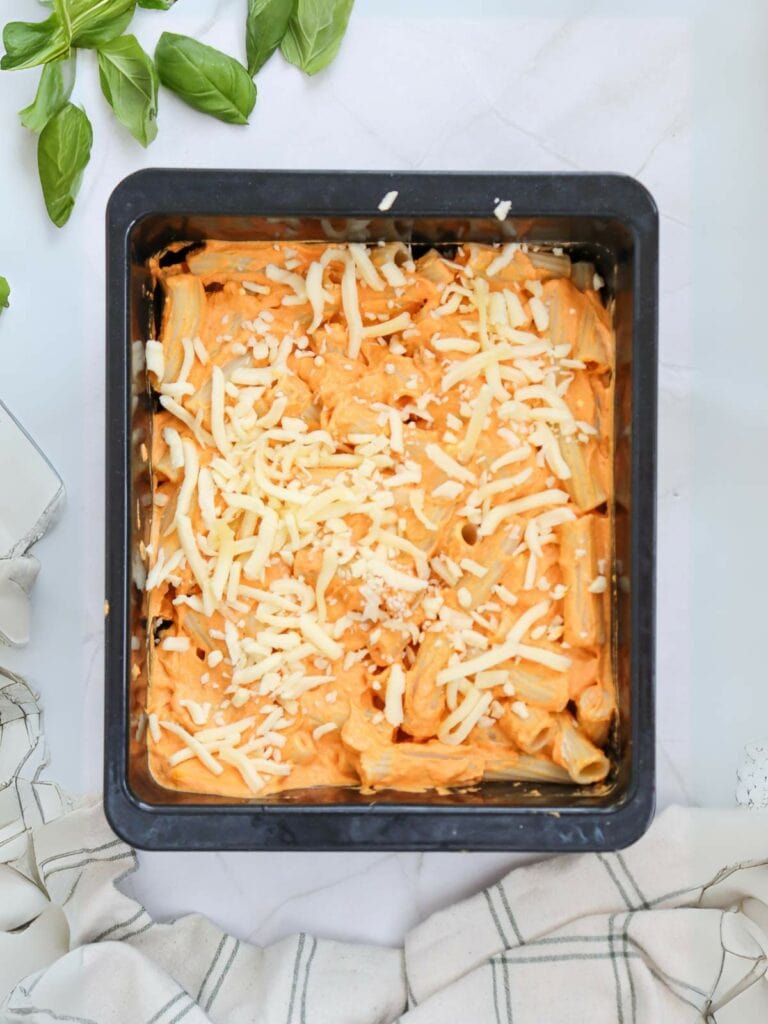 A square baking pan filled with uncooked pasta topped with cheese.