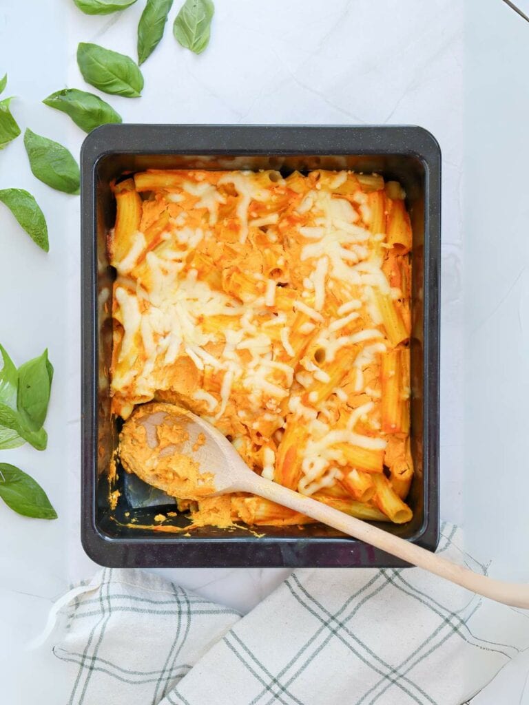 Baked pasta with melted cheese in a square pan, next to scattered basil leaves and a checkered cloth.