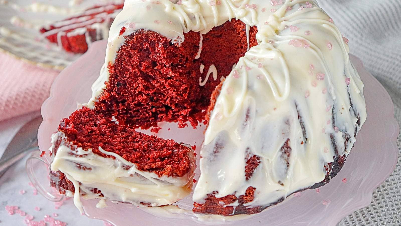 A ring-shaped red velvet cake with white icing is sliced to reveal its red interior, displayed on a pink cake stand.