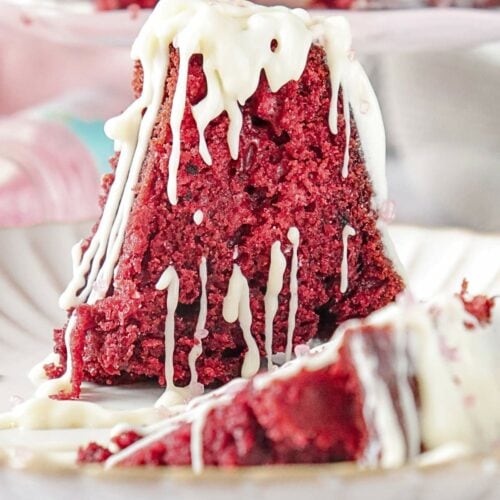 A slice of red velvet cake with white icing drizzles on a plate, with another plate of cake blurred in the background.