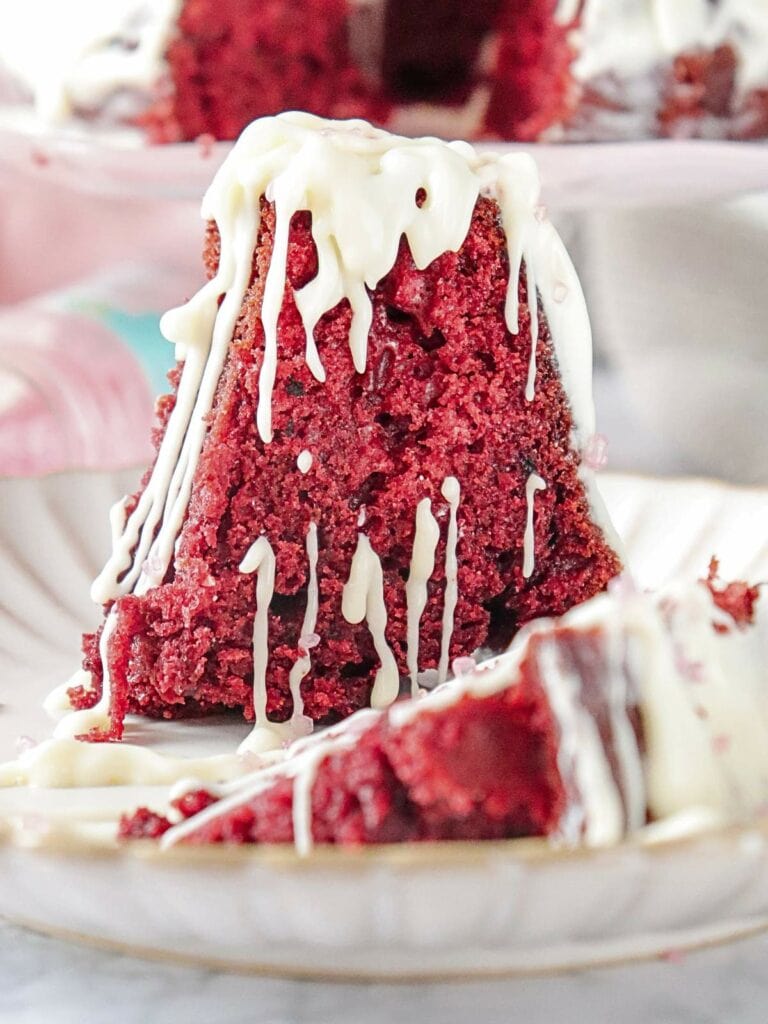 A slice of red velvet cake with white icing drizzles on a plate, with another plate of cake blurred in the background.