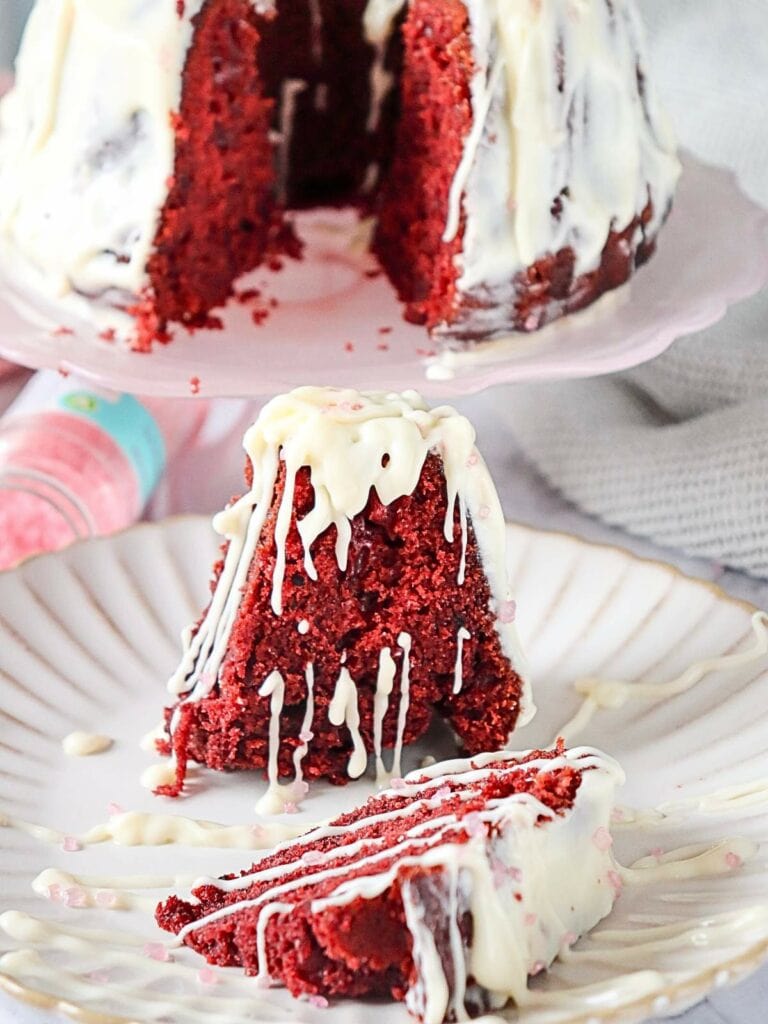 A slice of red velvet cake with white icing is on a plate, with the whole cake on a stand in the background.