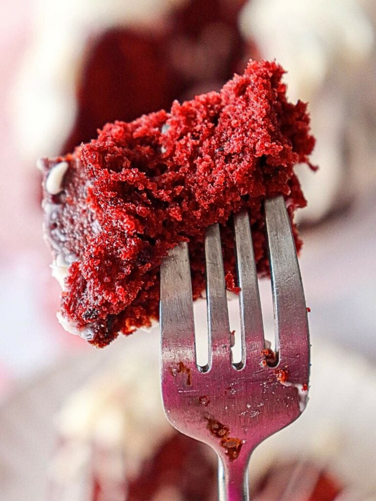 A fork holding a bite of red velvet cake with white frosting in the background.