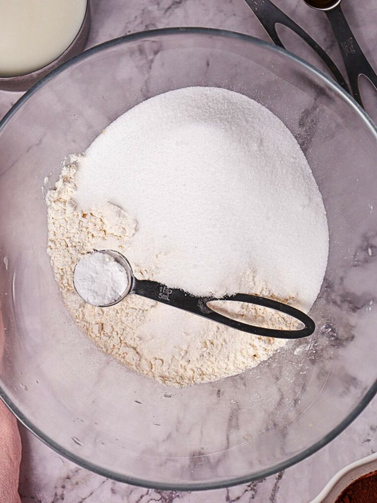 A glass bowl containing sugar and flour with a measuring spoon on a marble countertop.