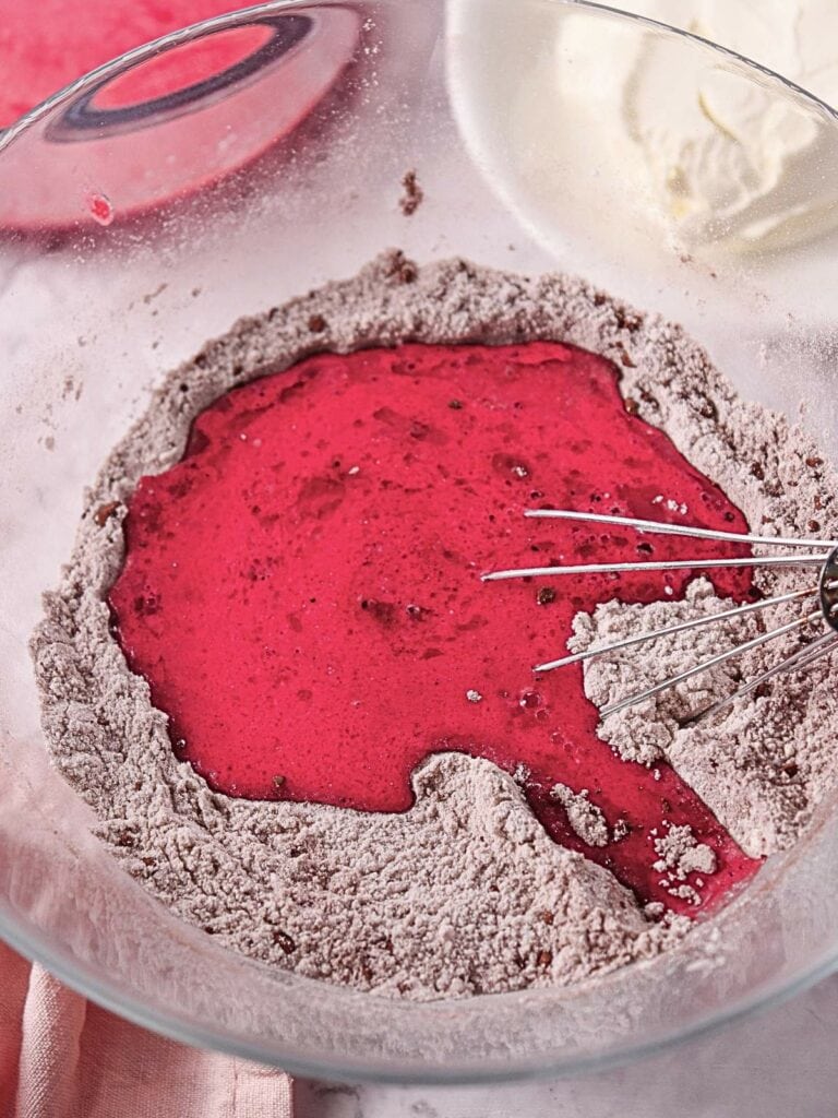 A glass bowl with dry cake mix and vibrant red liquid being whisked together.