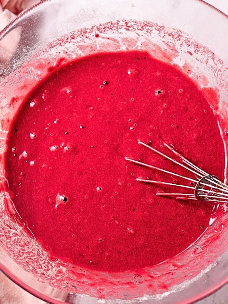 A glass bowl containing bright red batter with a whisk resting inside.