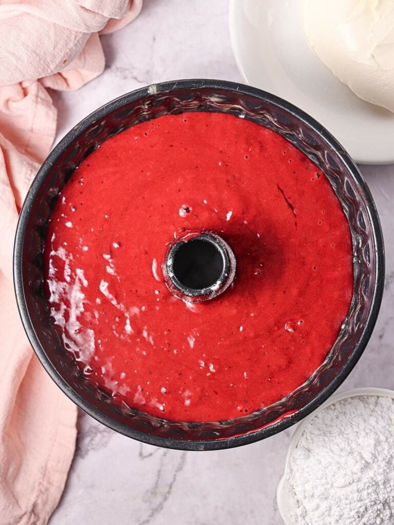 Red cake batter in a bundt pan on a marble surface, surrounded by a pink cloth, a bowl of white frosting, and a small bowl of flour.