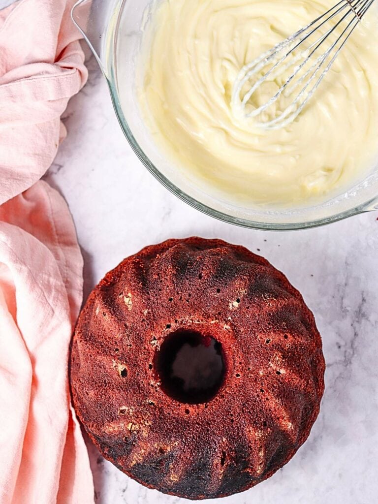 A bundt cake on a marble surface next to a mixing bowl of creamy batter with a whisk.