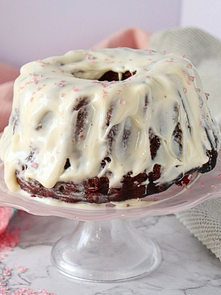 A chocolate bundt cake on a glass stand, covered with white icing and sprinkled with pink sugar.