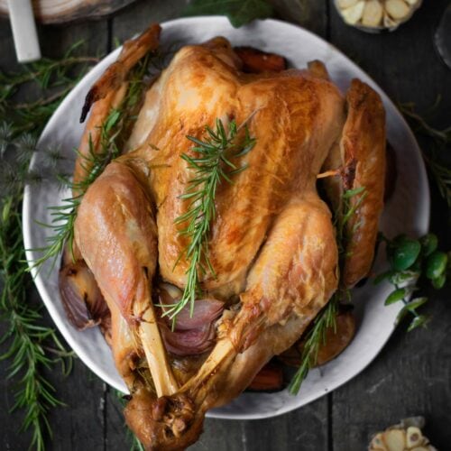 Roasted turkey on a plate, garnished with fresh rosemary, surrounded by garlic cloves and a glass of water on a dark wooden table.